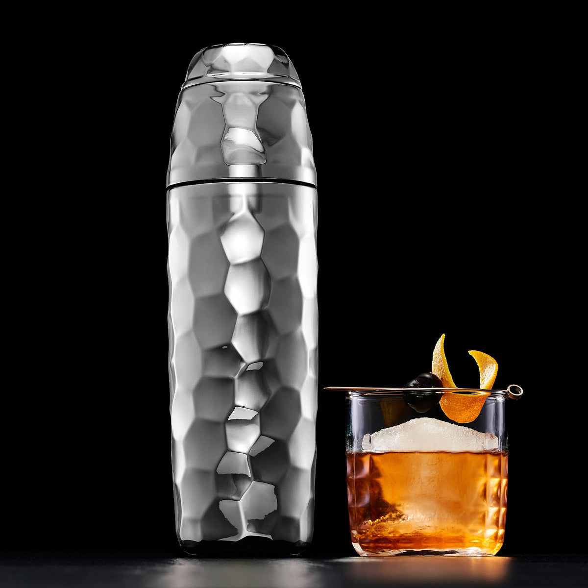 Chrome hexclad cocktail shaker next to a Negroni with ice and orange peel on a black background